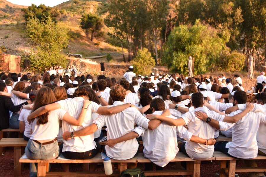 large group of teens in white shirts hanging onto eachothers shoulders while sitting down and watching a performance outside.