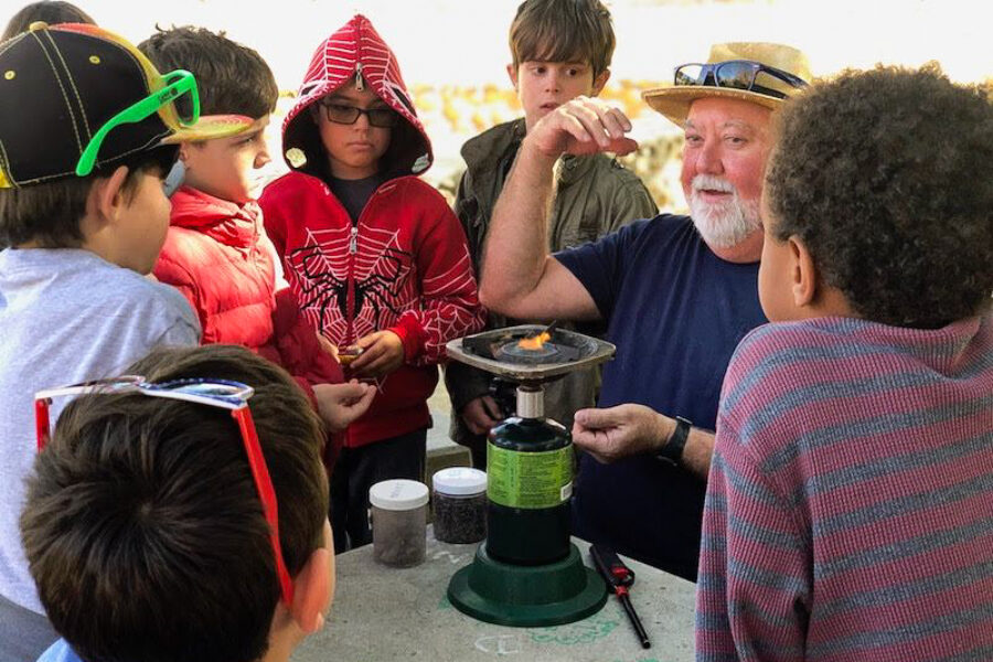 man demonstrating something with a camping burner to a group of kids outside.