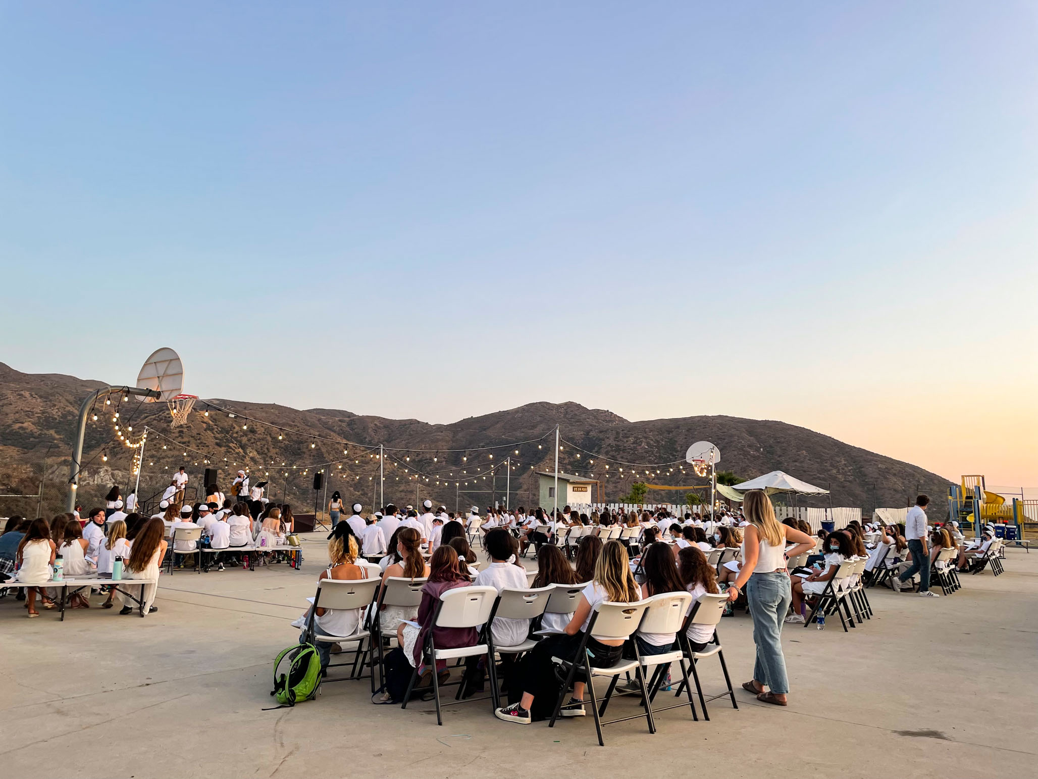 teens sitting in folding chairs outside watching a performance on a basketball court.