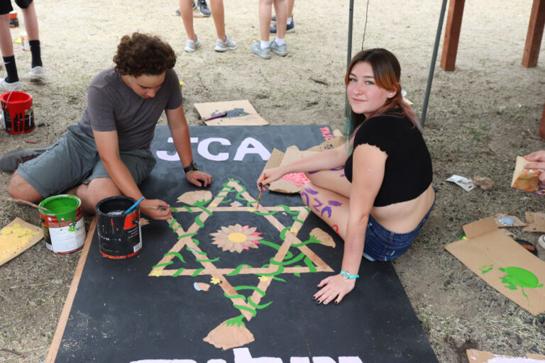two teens painting a sign on the ground that says JCA with a star of david.