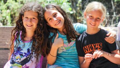 three girls sitting and smiling on a bench outside.