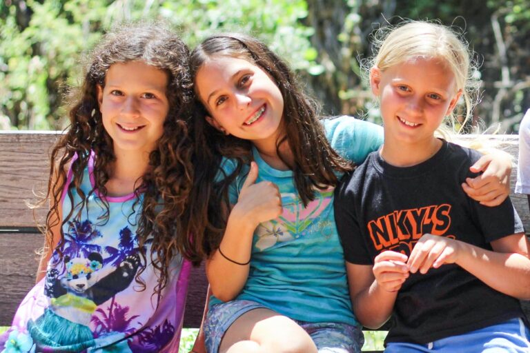 three girls sitting and smiling on a bench outside.